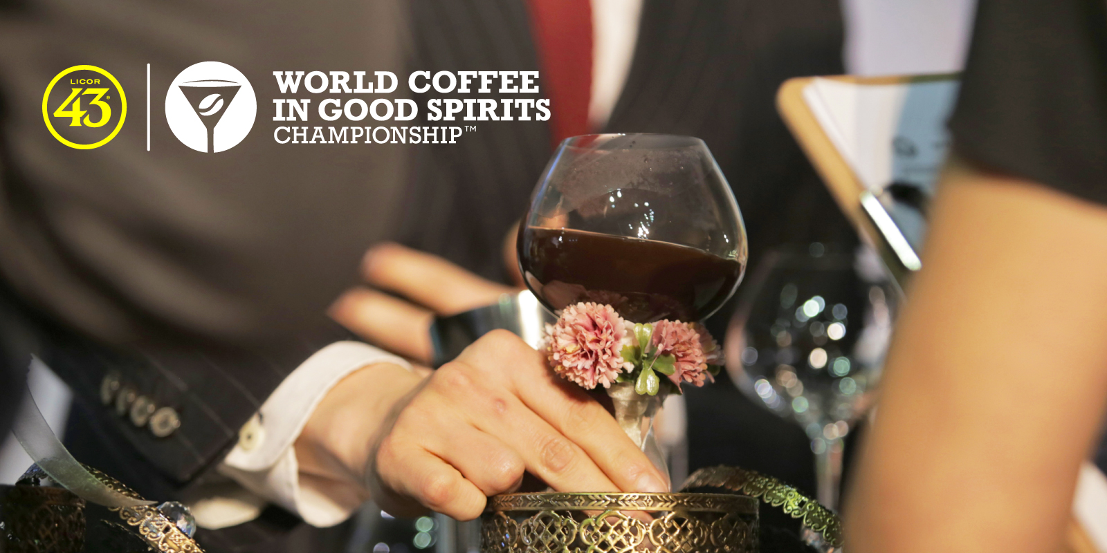 A squat piece of stemmed glassware decorated with dried flowers is served to a judge. There is a coffee cocktail in the cup. The image has the Licor 43 & World Coffee In Good Spirits Championships logos overlayed.